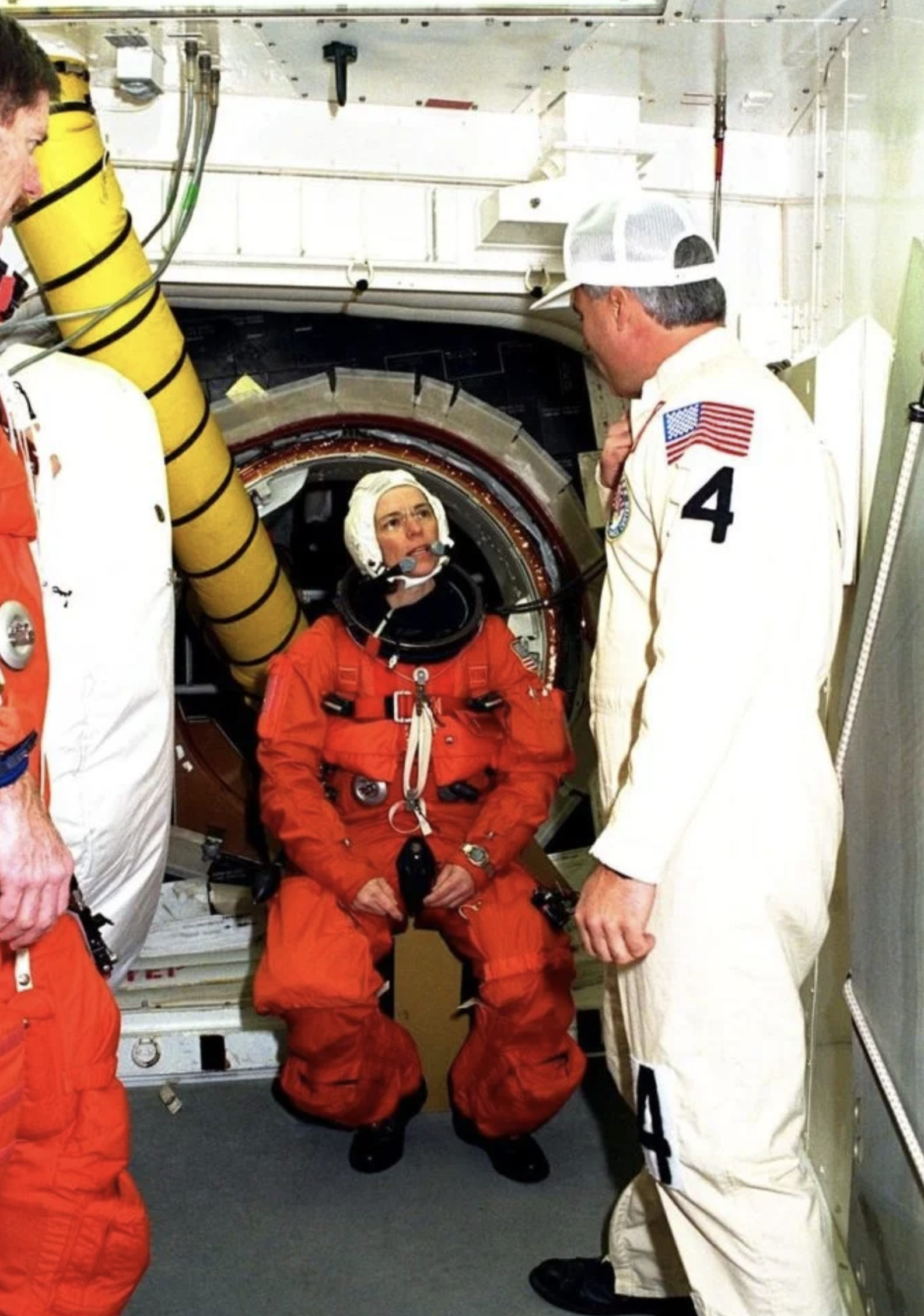 Female astronaut in a bulky orange spacesuit on a space shuttle talking to NASA techs in white form-fitting suits